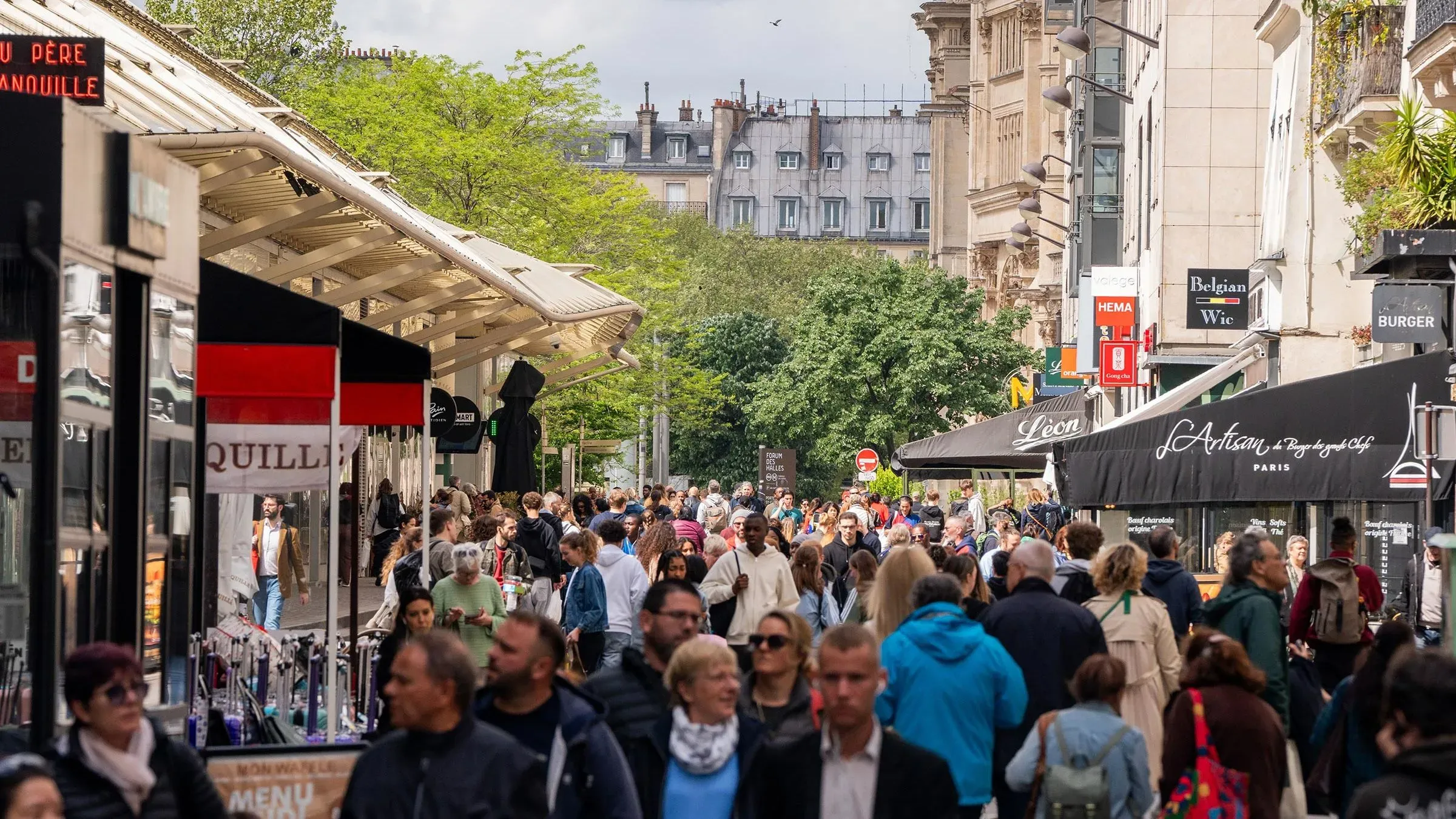 شارع مزدحم في مدينة باريس، حيث يتجمع العديد من الناس حول المحلات والمطاعم، مع أشجار خضراء في الخلفية.