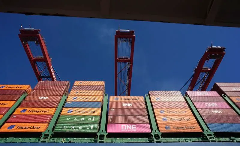 حاويات شحن ملونة في ميناء مع رافعات تحميل تحت سماء زرقاء، تعكس تأثير التعريفات الجمركية الأمريكية على التجارة العالمية.