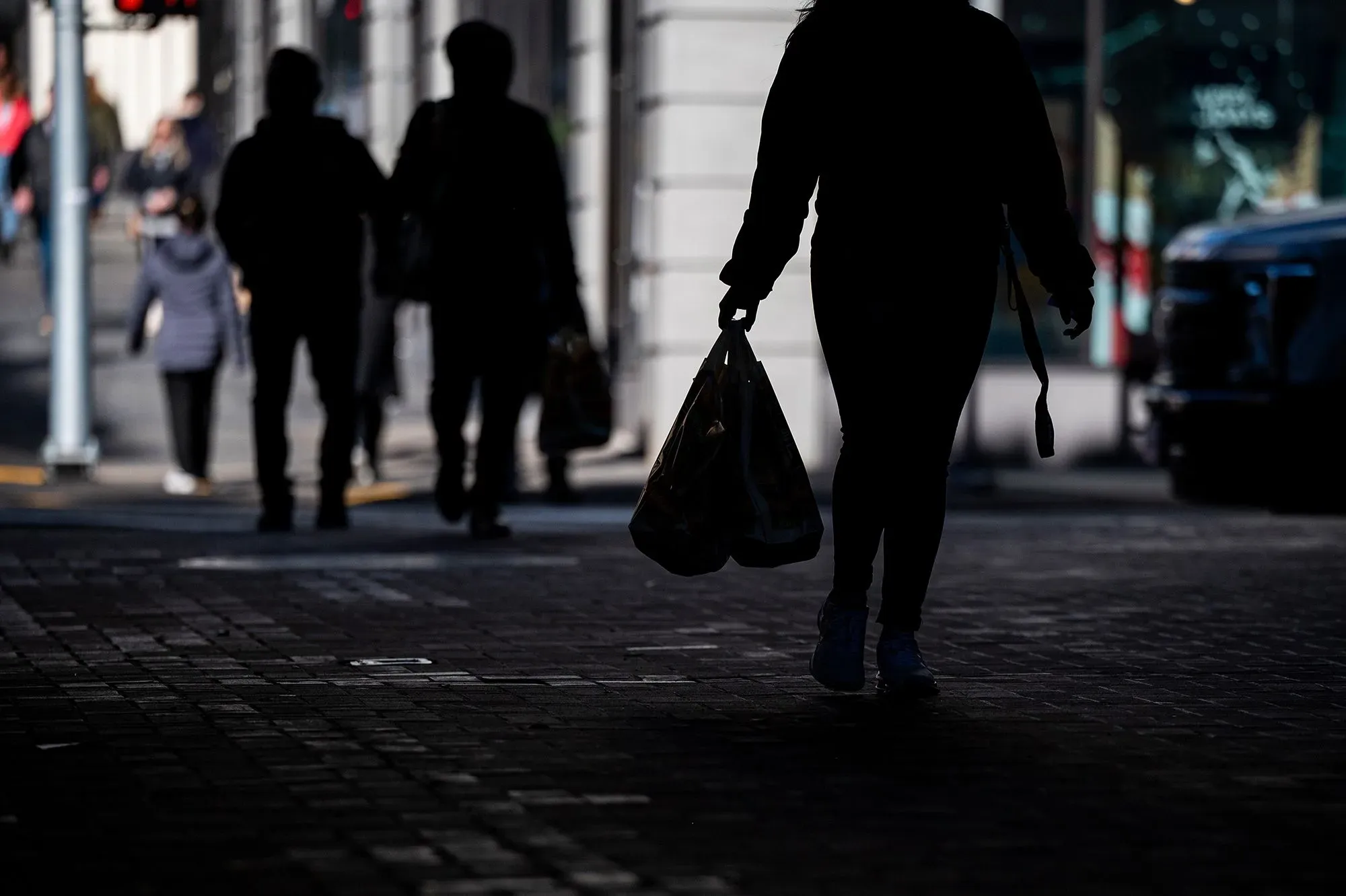 امرأة تحمل حقيبة تسوق تمشي في الشارع، بينما تظهر أشخاص آخرون في الخلفية. تعكس الصورة تأثير التعريفات الجمركية على الأسر الأمريكية.