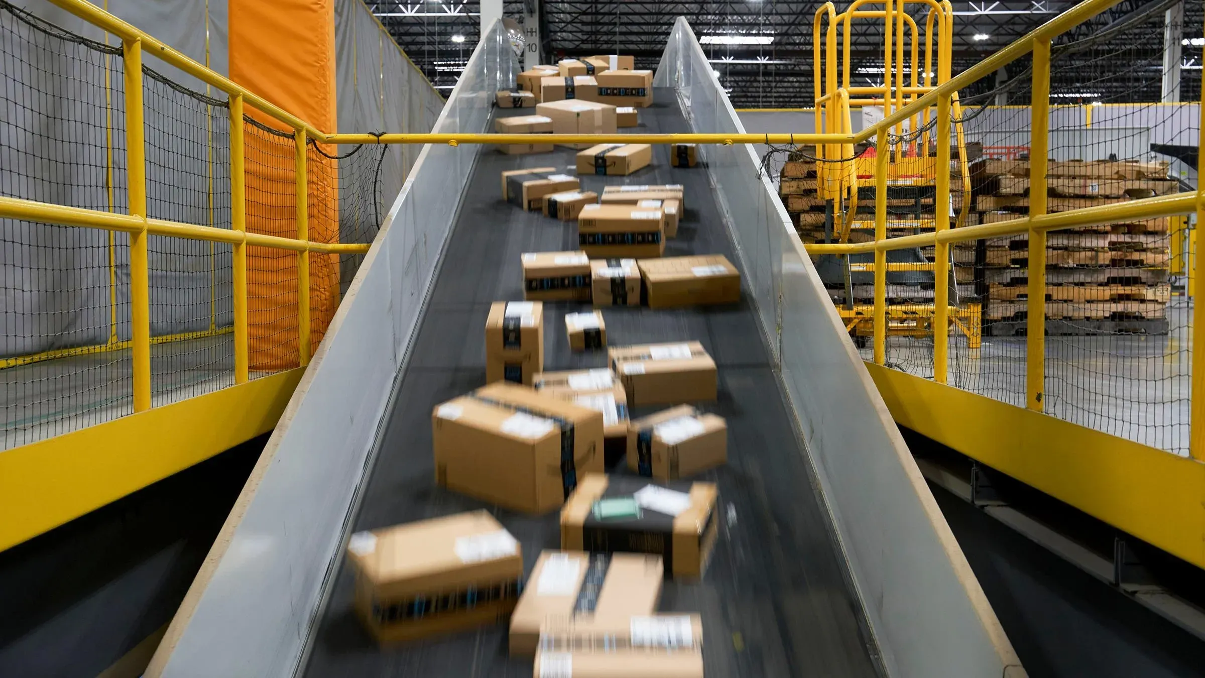 صناديق شحن تتدفق على حزام ناقل في مستودع أمازون، تعكس نشاط التجارة الإلكترونية وتأثير التعريفات الجمركية على الأسعار.