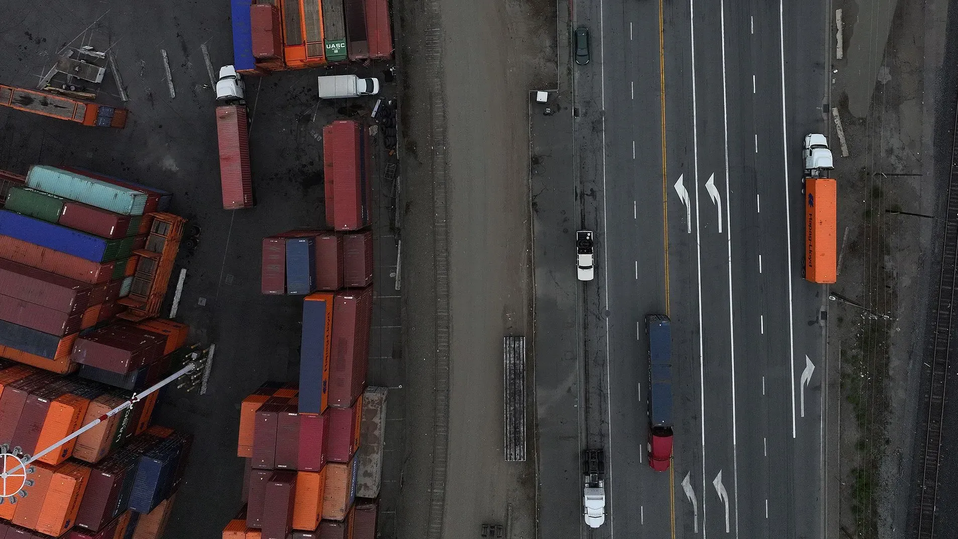 صورة جوية لميناء شحن يظهر حاويات ملونة مركونة بجانب الطريق، مع شاحنات تسير على الطريق السريع، تعكس حركة التجارة والاقتصاد.