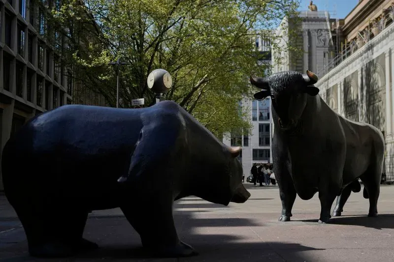 تمثالان لدب وثور يرمزان إلى تقلبات الأسواق المالية، مع خلفية لأشجار ومباني حضرية، مما يعكس الأجواء الاقتصادية الحالية.