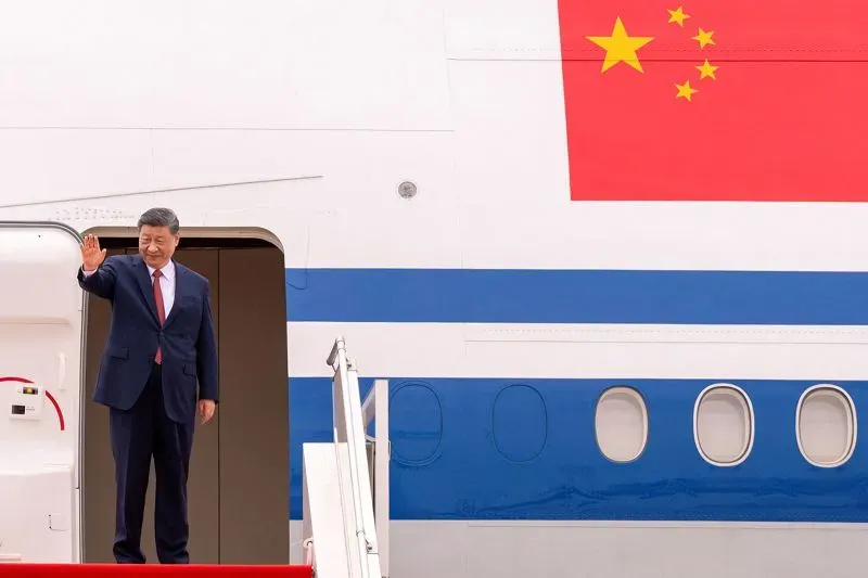 شي جين بينغ يلوح بيده أثناء مغادرته الطائرة، مع العلم الصيني واضح خلفه، مما يعكس موقف الصين في ظل التوترات التجارية مع الولايات المتحدة.