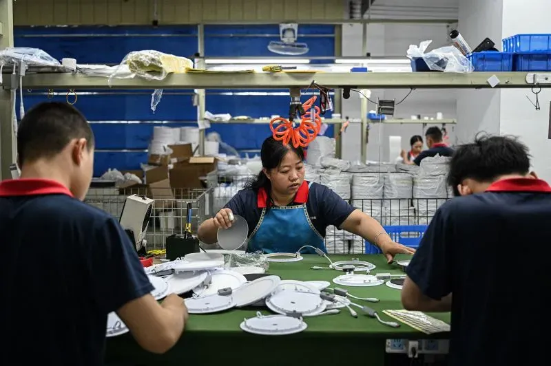 عمال في مصنع يقومون بتجميع المنتجات الكهربائية، مع التركيز على امرأة ترتدي مئزرًا أزرق وتعمل على خط الإنتاج.