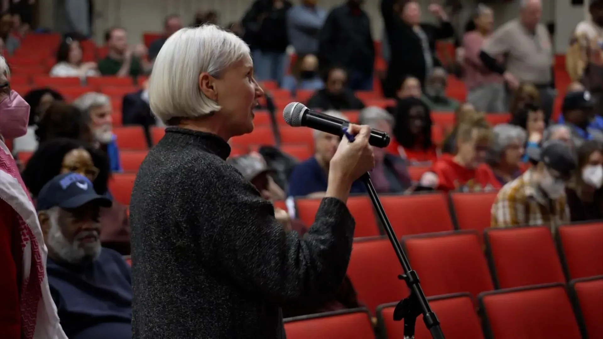 امرأة تتحدث إلى ميكروفون خلال اجتماع بلدي، مع جمهور متنوع خلفها، تعبر عن مشاعر الناخبين تجاه أداء المشرعين الديمقراطيين.