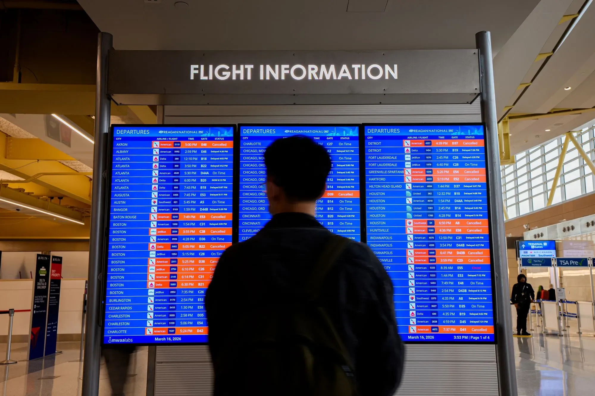 معلومات عن الرحلات في مطار مزدحم، حيث يظهر مسافر يتفقد تفاصيل الرحلات المغادرة، مع وجود تأخيرات وإلغاءات ملحوظة.