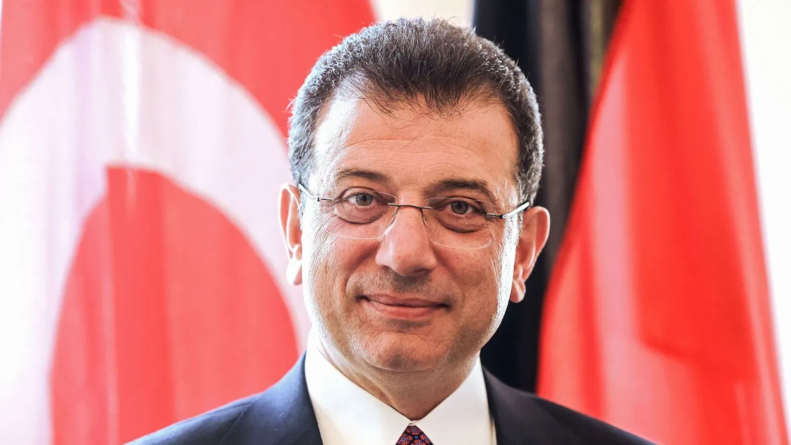 أكرم إمام أوغلو، رئيس بلدية إسطنبول، مبتسم أمام العلم التركي، بعد إدانته بتهمة إهانة موظف عام.