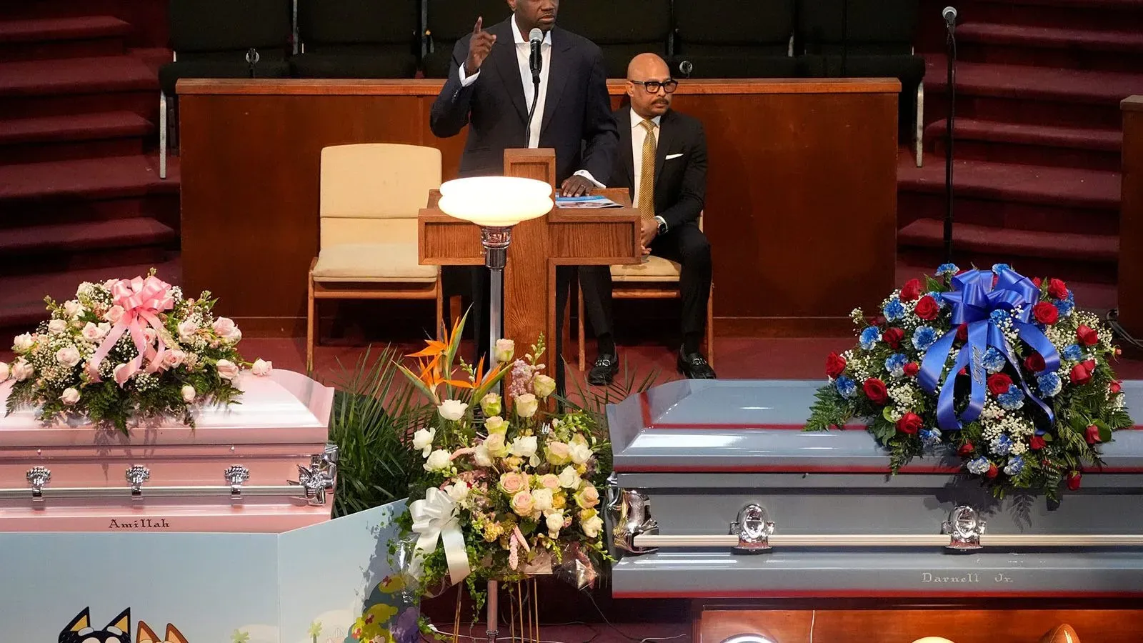 مراسم تأبين طفلين من ديترويت، مع تابوتين مزينين بالزهور، بينما يتحدث أحد الأشخاص في خلفية الكنيسة.
