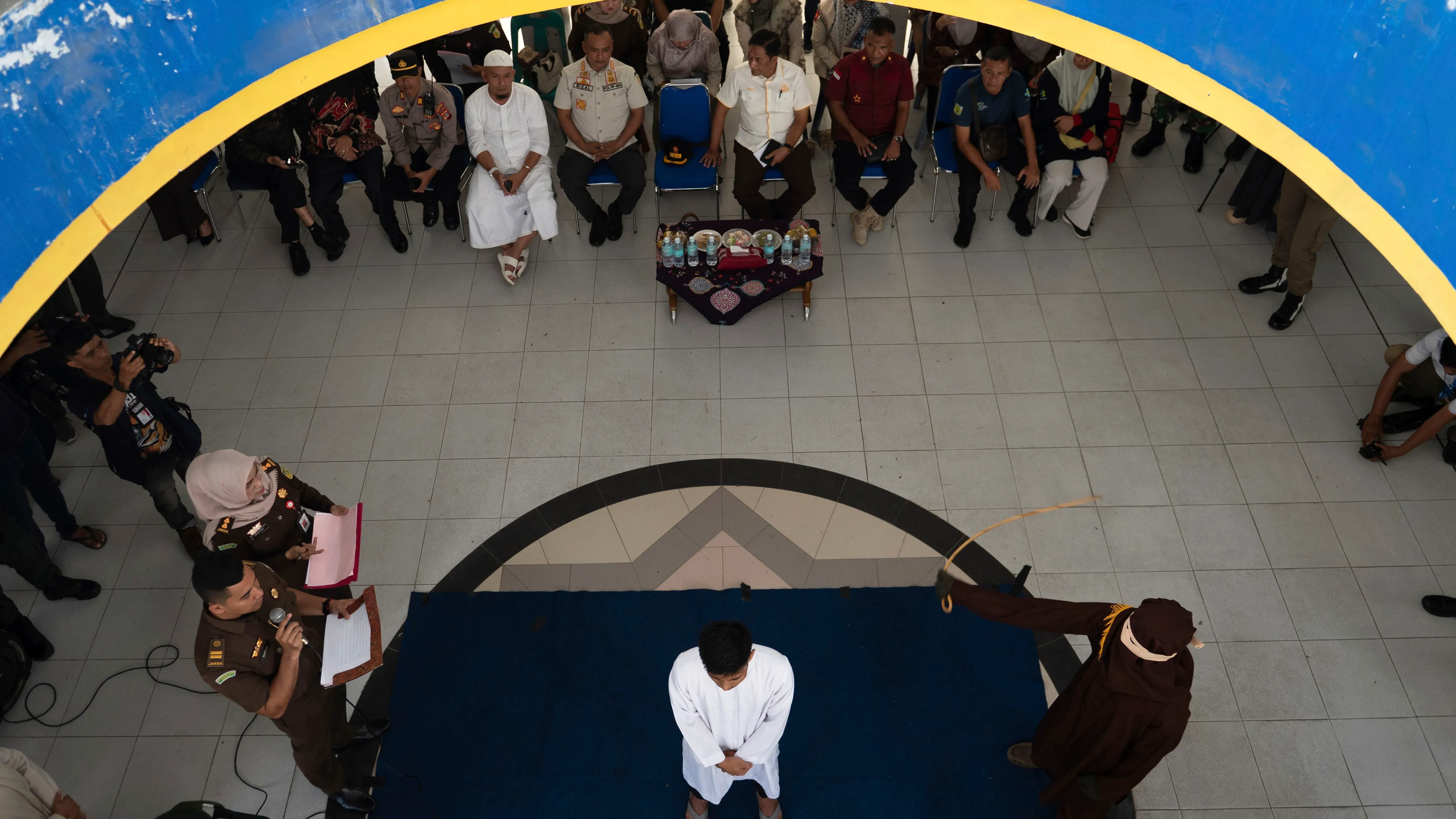 رجل يرتدي ملابس بيضاء يتعرض للجلد في قاعة عامة، بينما يشاهد الجمهور من حوله، في إطار تنفيذ عقوبة الشريعة الإسلامية في إندونيسيا.