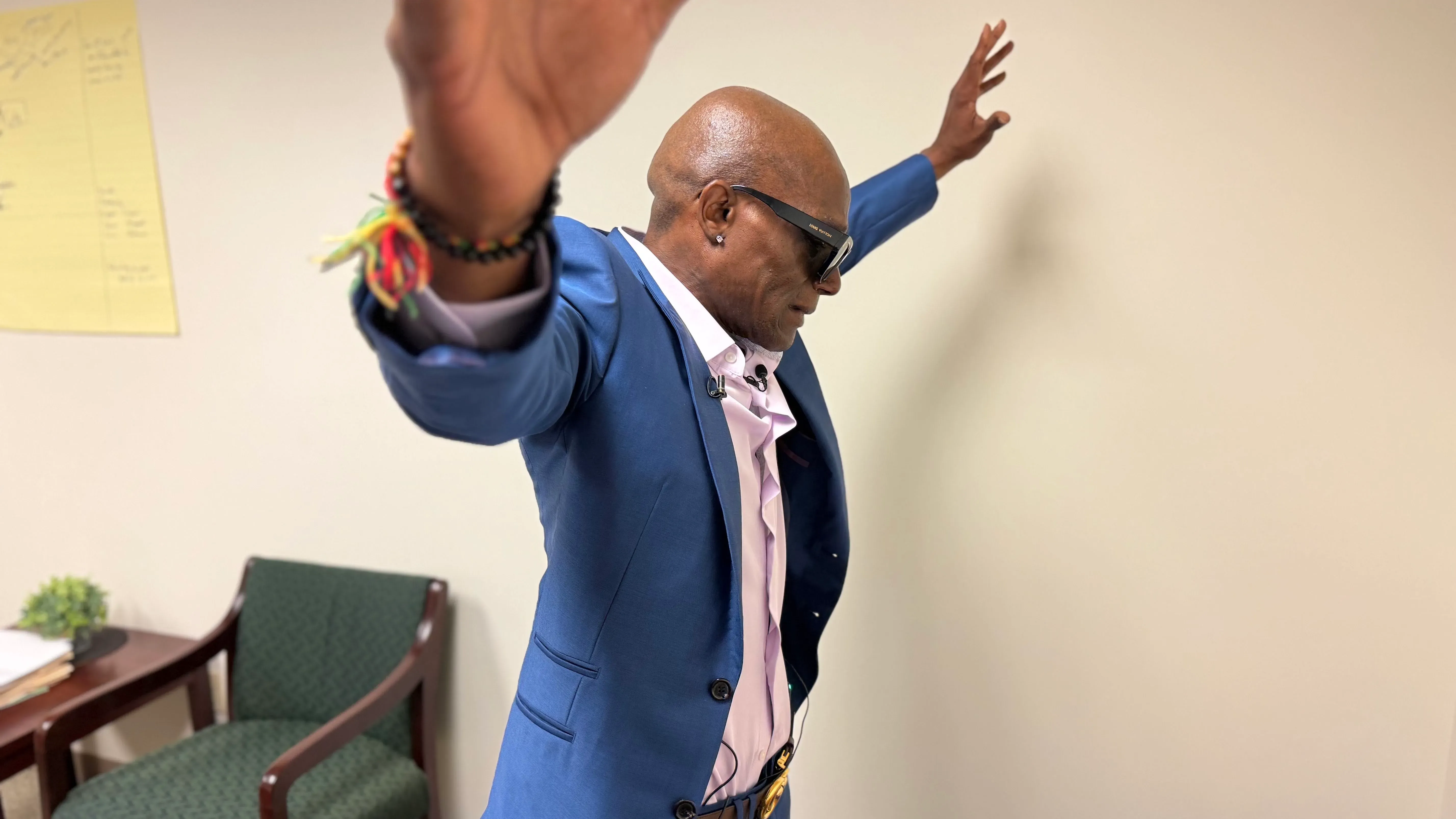 رجل يرتدي بدلة زرقاء وقميص وردي، يرفع ذراعيه في وضعية تعبيرية، في غرفة ذات جدران فاتحة. تعكس الصورة مشاعر الغضب والإحباط بعد تجربة الاعتقال.