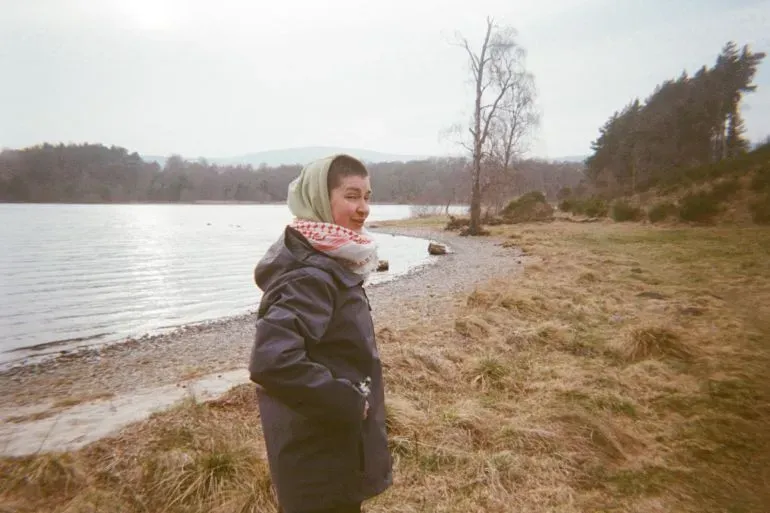 امرأة ترتدي حجابًا وتبتسم، تقف على ضفة بحيرة محاطة بالطبيعة، تعكس روح الأمل في سياق الإضراب عن الطعام.