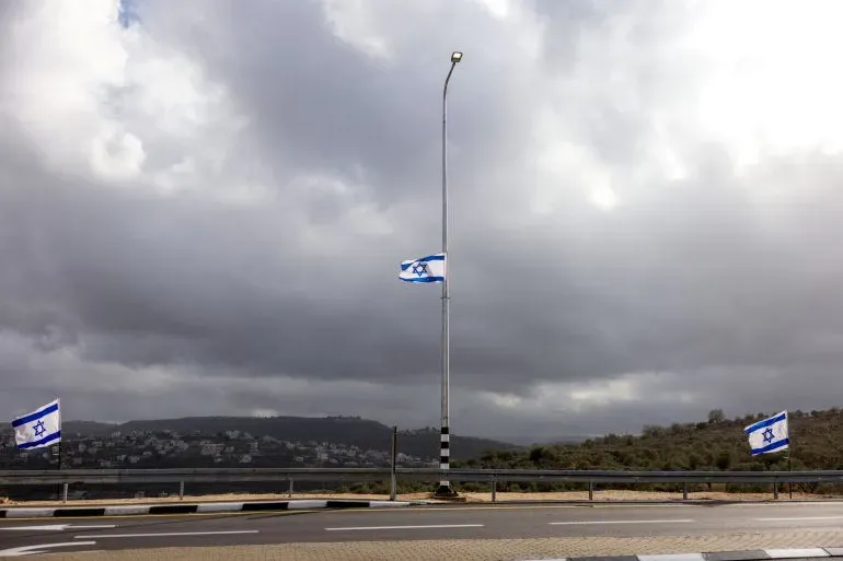 علم إسرائيلي يرفرف على عمود إنارة في منطقة جبلية تحت سماء غائمة، مع مشهد لقرية في الخلفية.