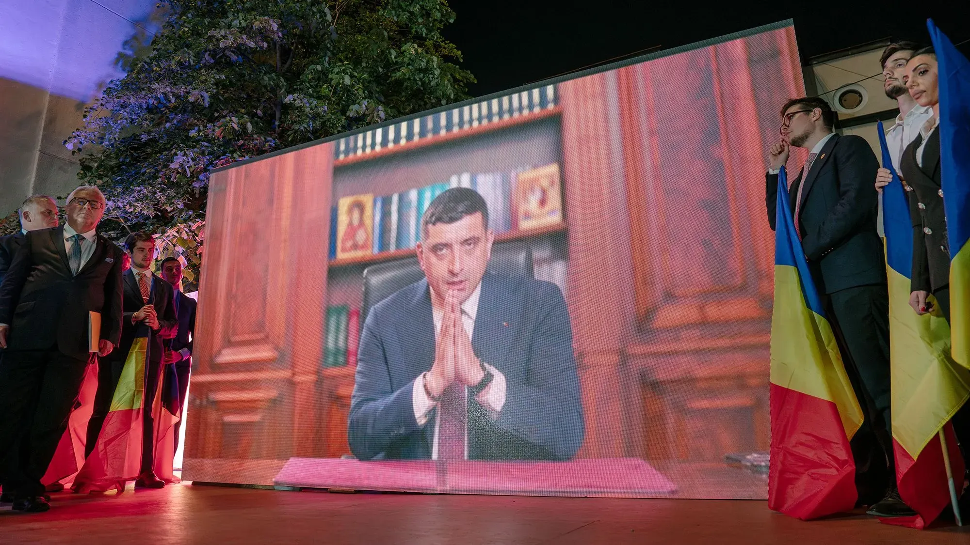 رجل يظهر على شاشة كبيرة خلال حدث سياسي في رومانيا، بينما يقف بجانبه مجموعة من الأشخاص يحملون الأعلام الرومانية.