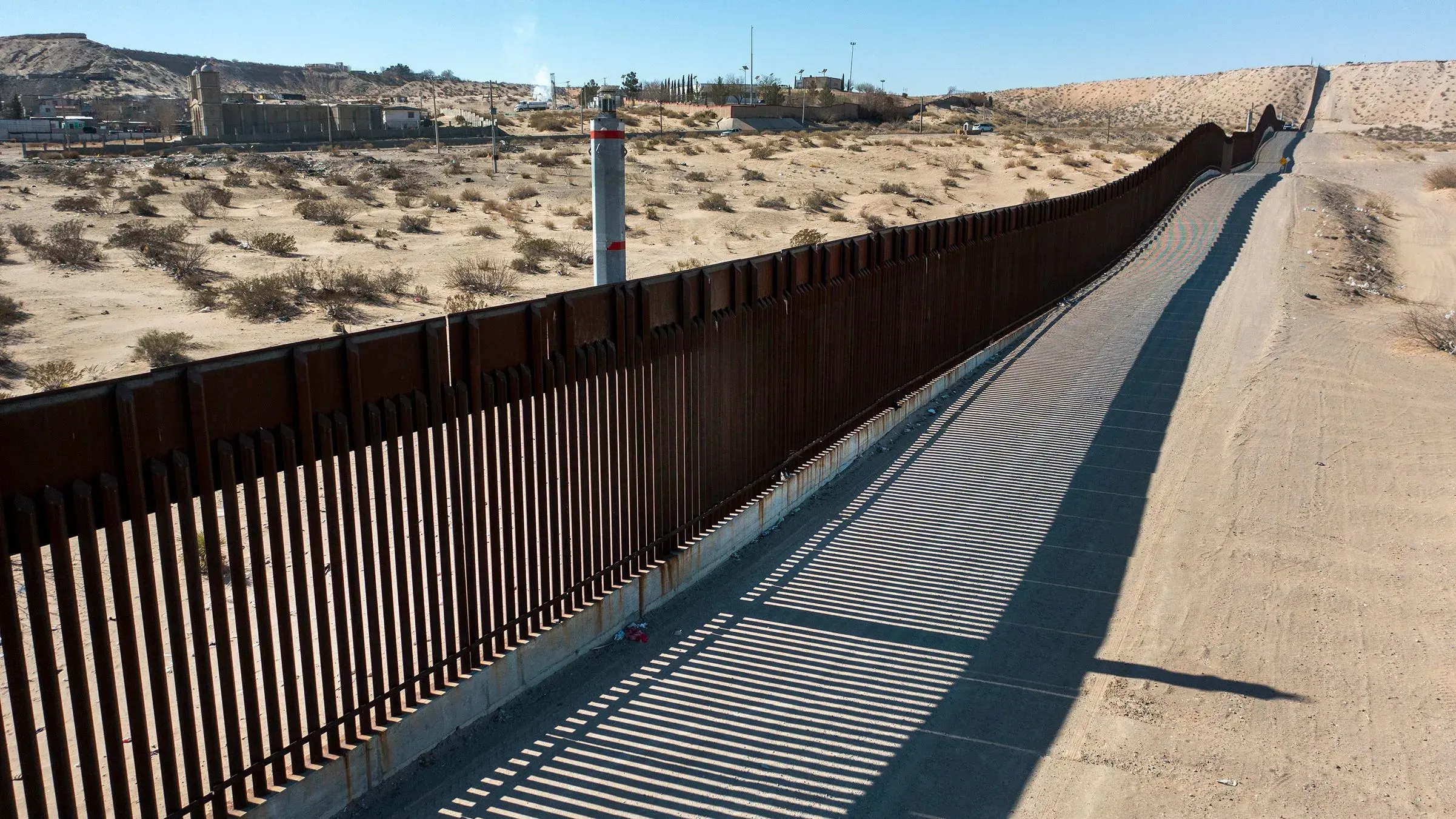 سياج الحدودي بين الولايات المتحدة والمكسيك في منطقة صحراوية، مع تفاصيل عن الطريق المجاور وظلاله، يعكس جهود تأمين الحدود الجنوبية.