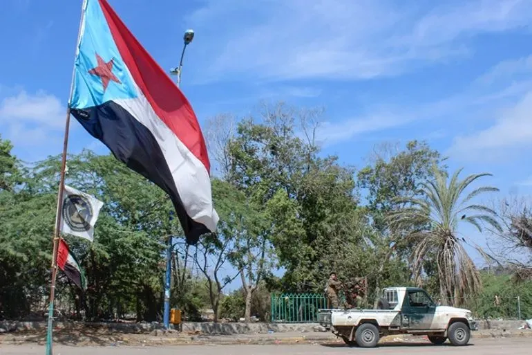 علم المجلس الانتقالي الجنوبي يرفرف في عدن، مع وجود سيارة عسكرية في الخلفية، مما يعكس التوترات السياسية والأمنية في اليمن.