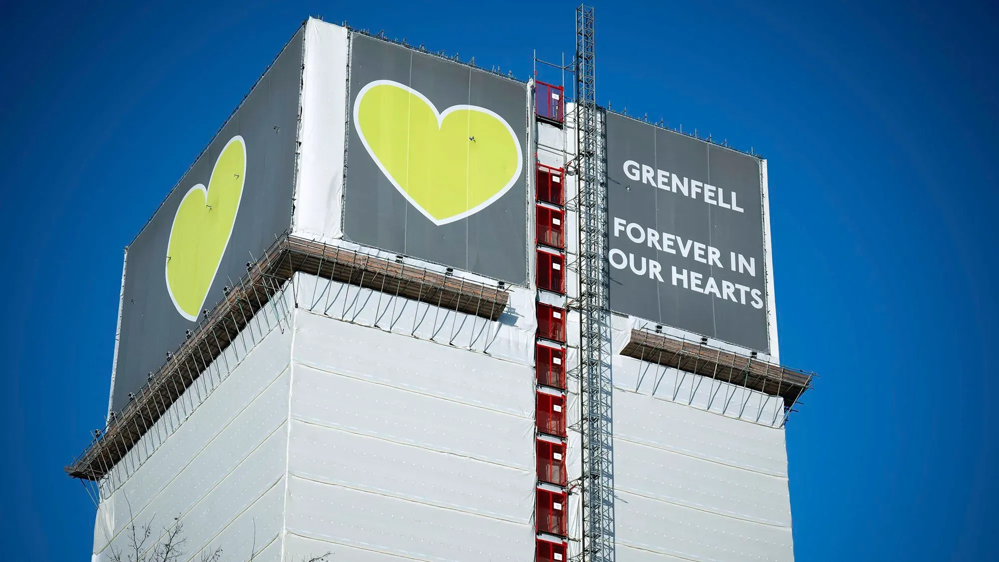 برج غرينفيل في لندن مغطى بشبكة بيضاء، مع لافتة تحمل عبارة "غرينفيل في قلوبنا إلى الأبد" وصورة قلوب صفراء، مما يرمز إلى الذكرى.