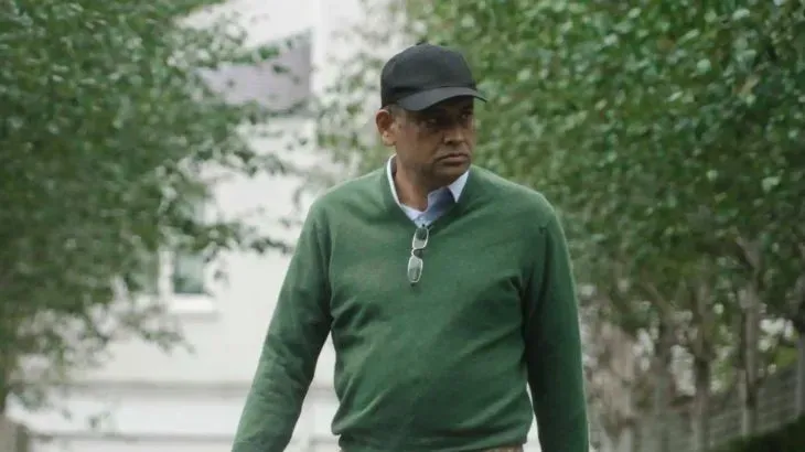 سيف الزمان شودري، وزير الأراضي البنغلاديشي السابق، يتجول في حي بلندن، وسط أشجار، مع نظارات وقبعة.