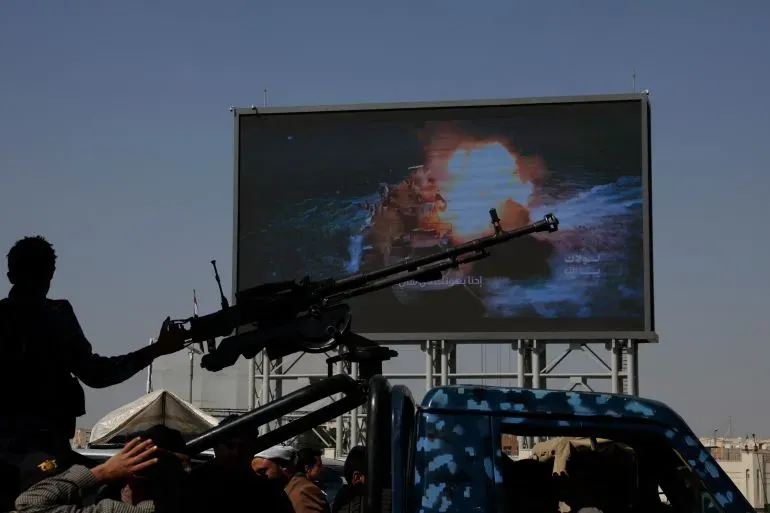 مشهد يظهر شخصًا يحمل سلاحًا مضادًا للطائرات أمام شاشة كبيرة تعرض صورة لحريق في سفينة، في سياق الهجمات الحوثية على السفن.