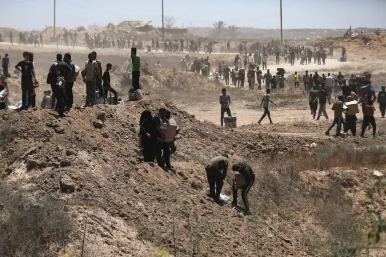 مواطنون فلسطينيون يجمعون المساعدات الإنسانية في غزة، مع وجود حشود في الخلفية، وسط ظروف صعبة بعد النزاع.