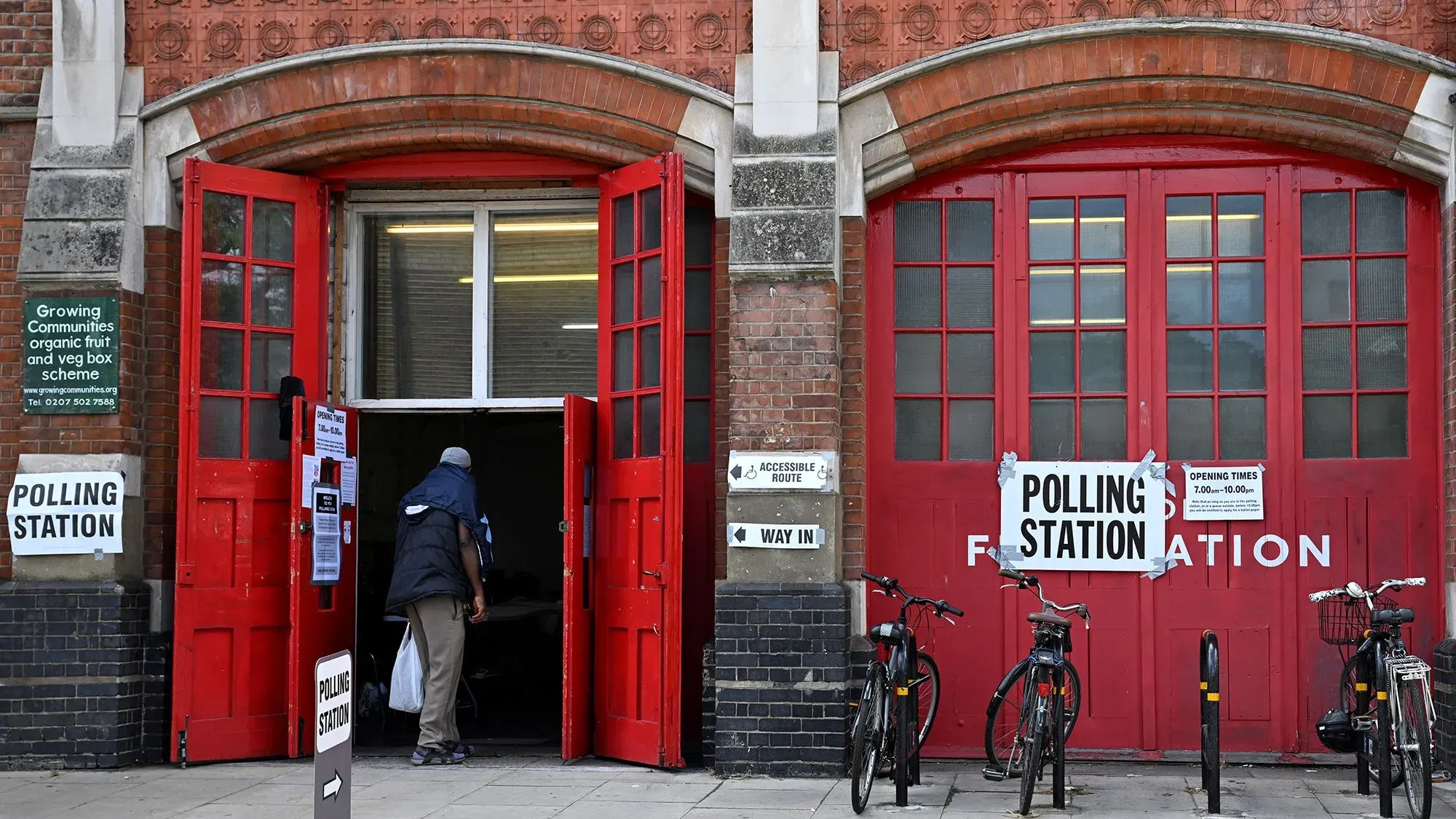 مدخل مركز الاقتراع مع أبواب حمراء، حيث يدخل ناخب، مع وجود لافتات توضح مواعيد العمل وإمكانية الوصول، ودراجات متوقفة بالخارج.