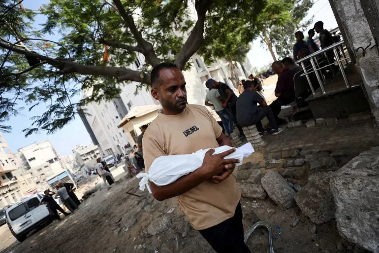 رجل يحمل طفلاً مغطى ببطانية بيضاء في غزة، وسط أجواء حزينة تعكس معاناة الأطفال بسبب المجاعة والنزاع المستمر.