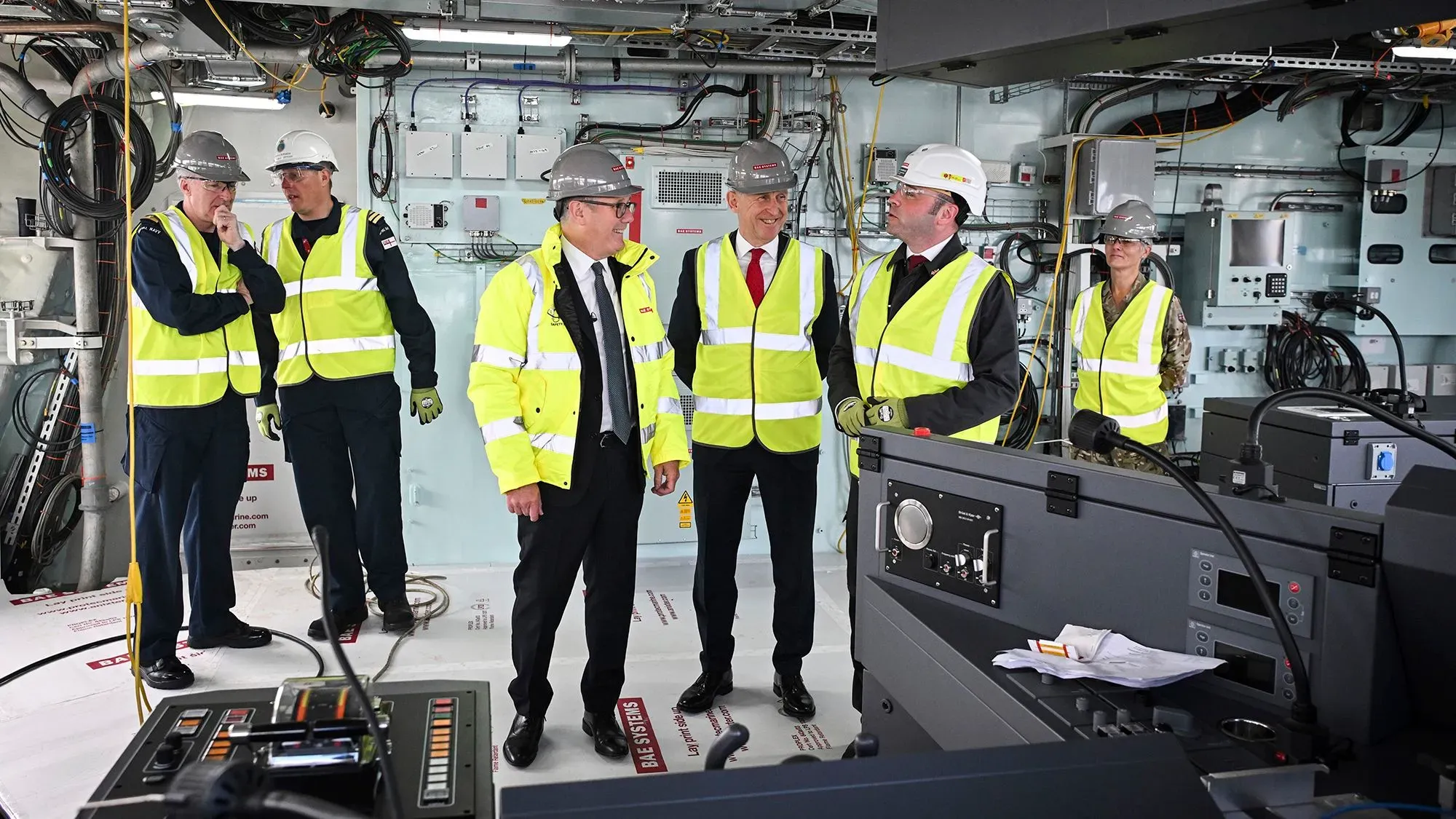 مجموعة من المسؤولين يرتدون سترات عاكسة وخوذات، يتحدثون في داخل غواصة جديدة قيد الإنشاء، مع وجود معدات بحرية خلفهم.