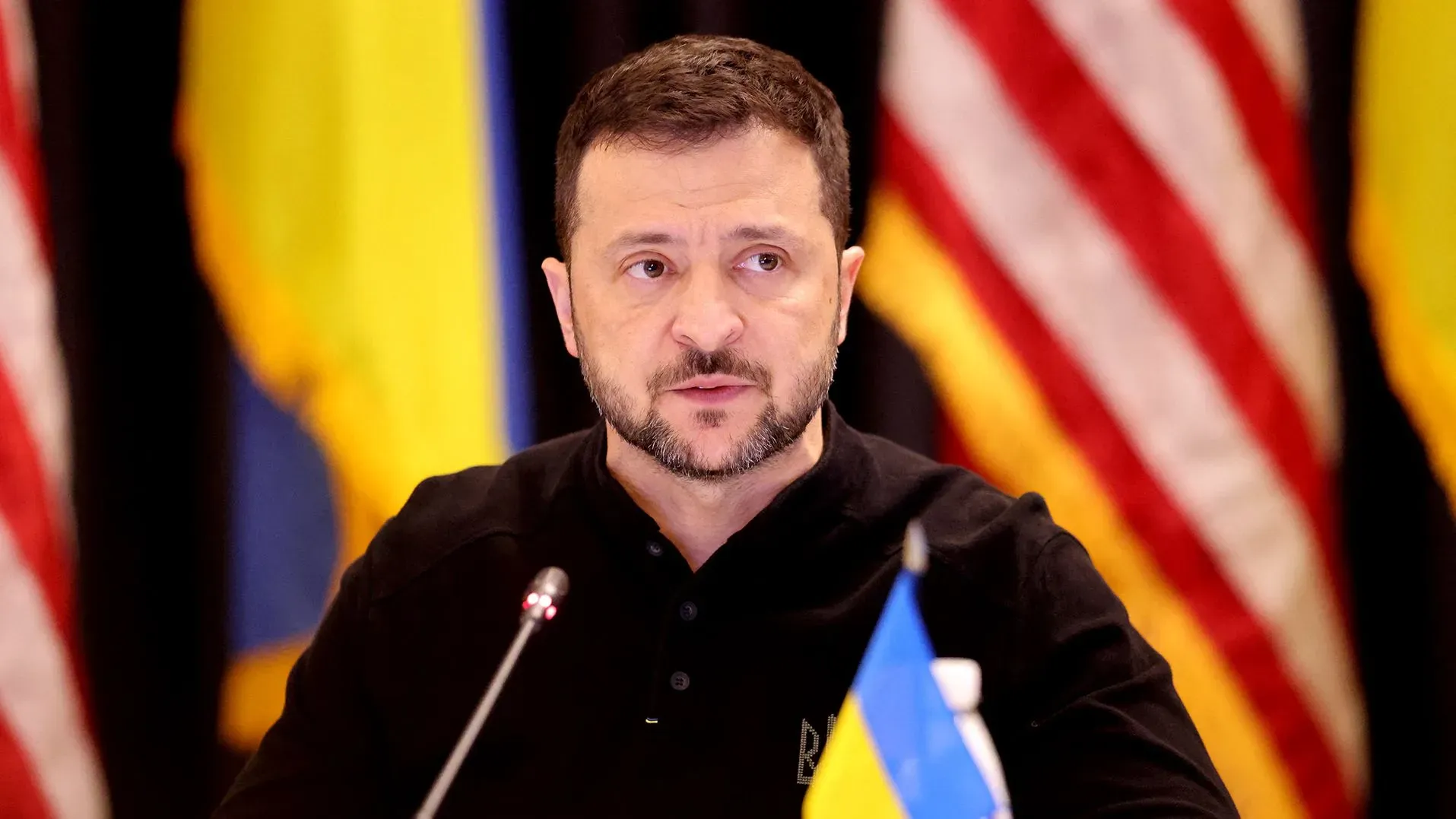 الرئيس الأوكراني فولوديمير زيلينسكي يتحدث خلال مؤتمر صحفي، مع العلم الأوكراني وخلفية الأعلام الأمريكية، مستعرضًا مصالح أوكرانيا في زمن الحرب.