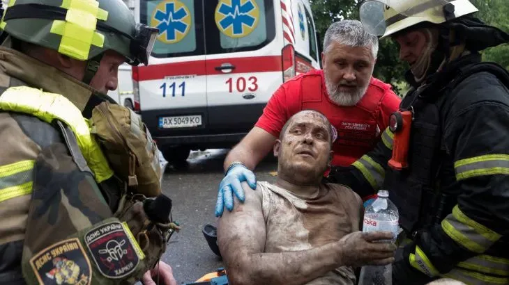 رجل مصاب يجلس على الأرض، يتلقى المساعدة من رجال الإنقاذ، بينما تظهر سيارة إسعاف في الخلفية. تعكس الصورة آثار الصراع في أوكرانيا.