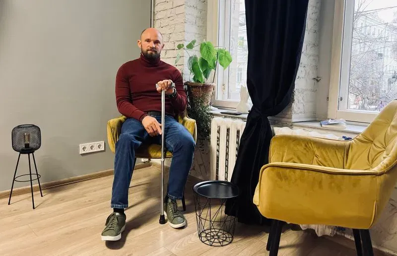 رجل يجلس على كرسي في غرفة مضاءة جيدًا، ممسكًا بعصا مساعدة، مع نباتات وزوايا مريحة، معبرًا عن تحديات إعادة التأهيل بعد الإصابة.