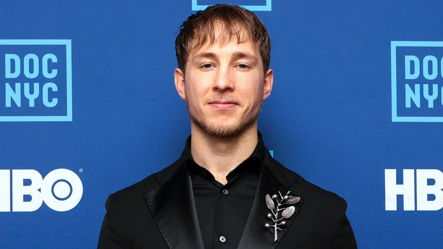 رجل يرتدي بدلة سوداء مع دبوس زينة، يقف أمام خلفية زرقاء تحمل شعارات \"DOC NYC\" و\"HBO\"، يبدو مبتسمًا وواثقًا.