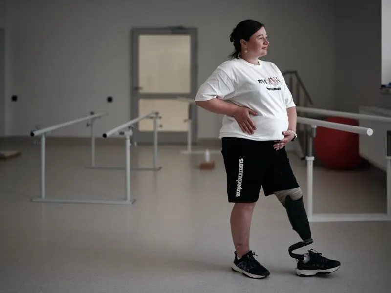 امرأة ترتدي ملابس رياضية وتستند إلى حائط في مركز إعادة تأهيل، تظهر بترًا في ساقها، تعكس التحديات التي تواجهها في التعافي بعد الإصابة.