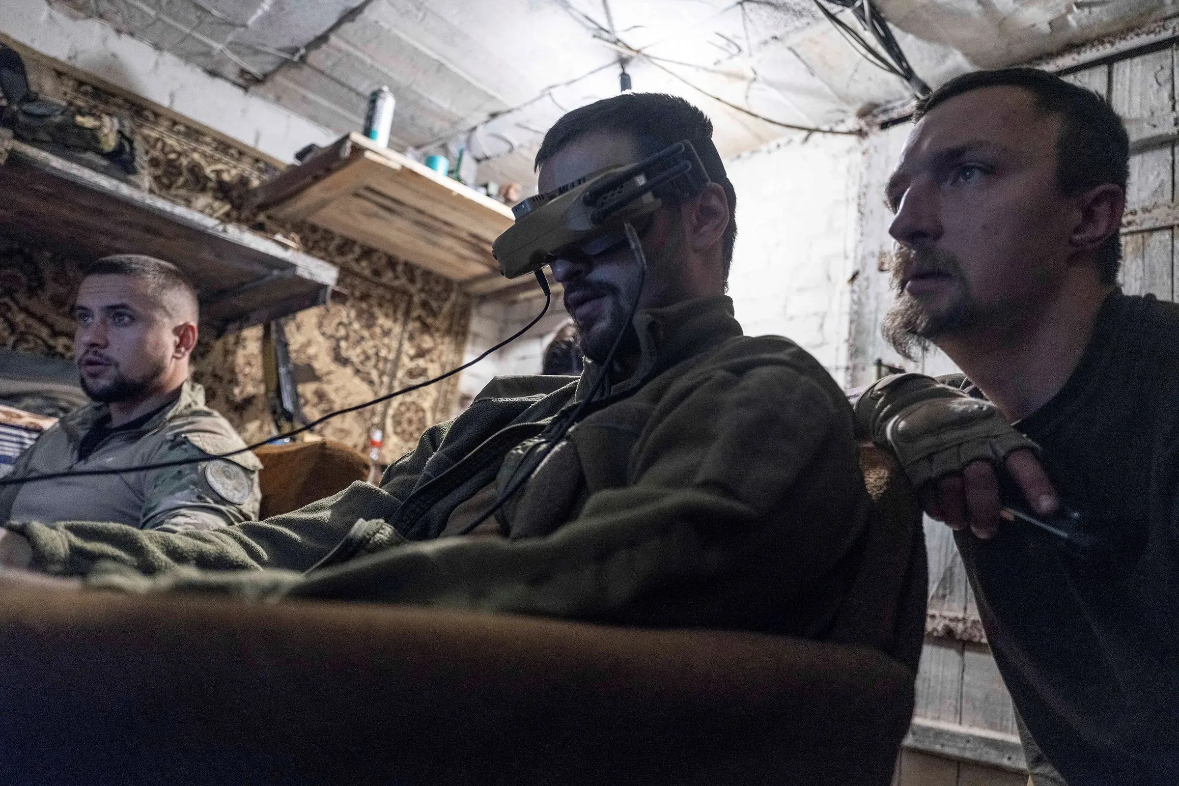 ثلاثة جنود أوكرانيين يجلسون في غرفة مظلمة، يراقبون شاشات بأجهزة تحكم، محاطين بأدوات عسكرية، في سياق الحرب الحديثة.