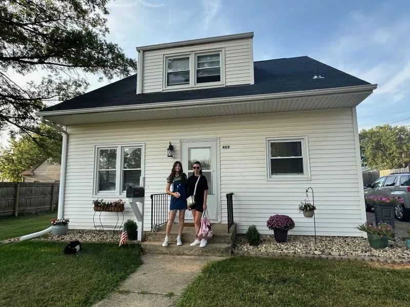 امرأتان أوكرانيتان واقفتان أمام منزل أبيض في هارتسفيل، حيث تعكسان تحديات إعادة بناء حياتهما بعد الهجرة إلى الولايات المتحدة.