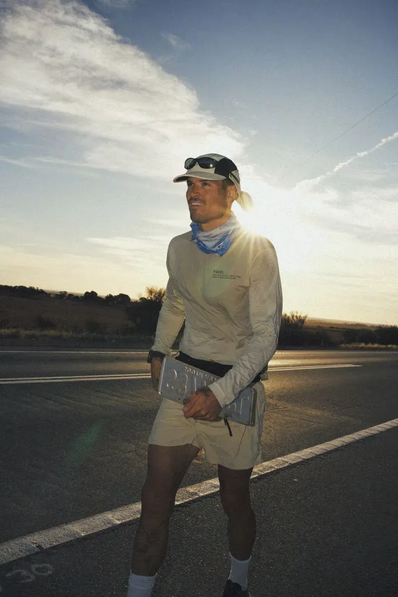 عداء يتقدم على طريق مفتوح تحت سماء زرقاء، مبتسمًا بينما يحمل زجاجة ماء، في إطار يجسد تحديات الجري لمسافات طويلة.