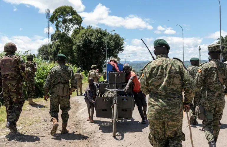 جنود من القوات الأوغندية يسيرون في منطقة ريفية في جمهورية الكونغو الديمقراطية، بينما يحمل مدنيون عربات محملة.