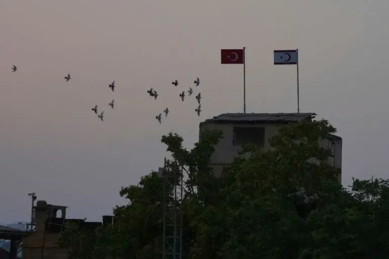 علمان يرفرفان فوق مبنى في قبرص، أحدهما علم تركيا والآخر علم جمهورية قبرص التركية، مع طيور تحلق في السماء.