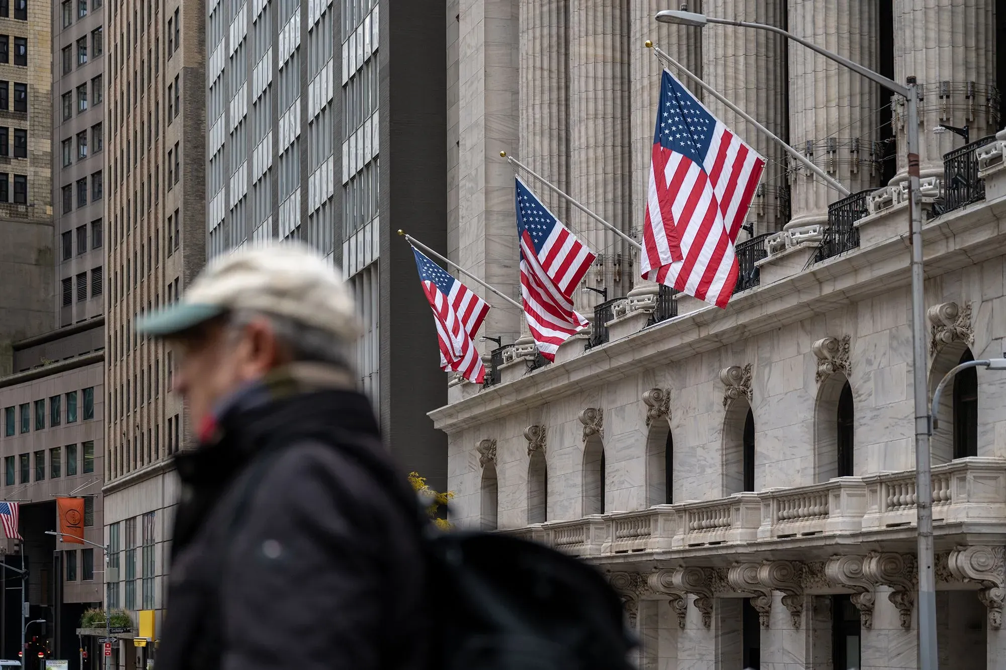 رجل يسير بجوار مبنى البورصة الأمريكية، مع أعلام الولايات المتحدة ترفرف في الهواء، مما يعكس النشاط المالي والاقتصادي.