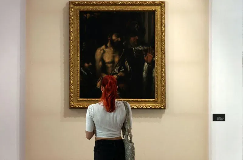 امرأة ذات شعر أحمر تقف أمام لوحة زيتية لتيتيان تُظهر مشهدًا دينيًا، في معرض فني بمدينة ليماسول.