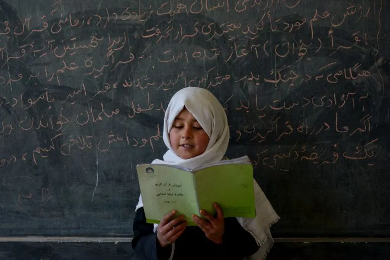 فتاة أفغانية ترتدي الحجاب تقرأ من كتاب أمام سبورة مكتوبة باللغة الفارسية، تعبير عن حرمان الفتيات من التعليم الثانوي في أفغانستان.