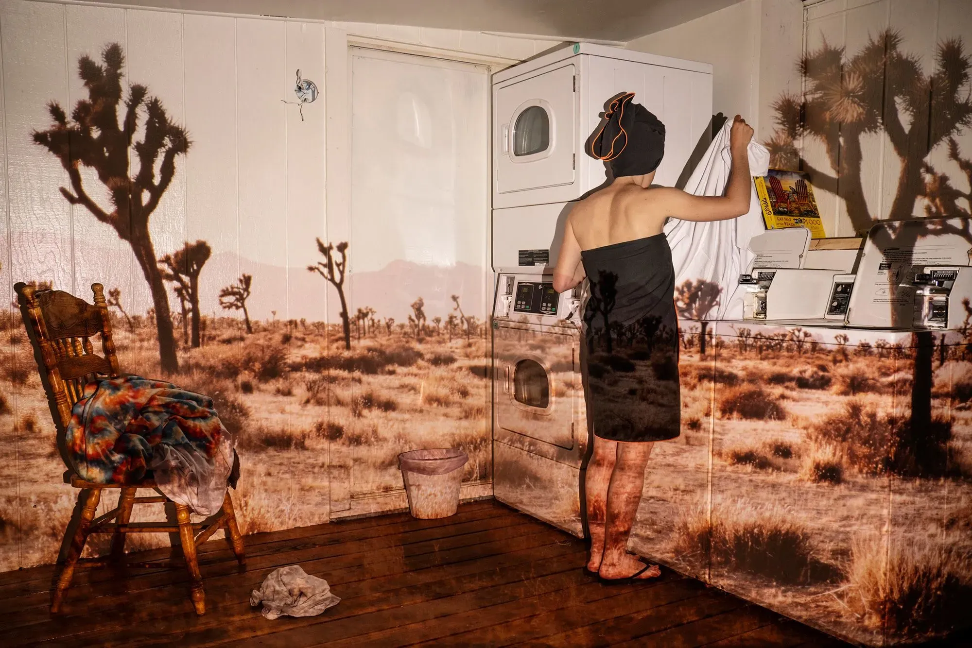 امرأة ترتدي منشفة تقف أمام غسالة في غرفة غسيل، بينما تظهر صورة لصحراء نيفادا على الجدران المحيطة بها.