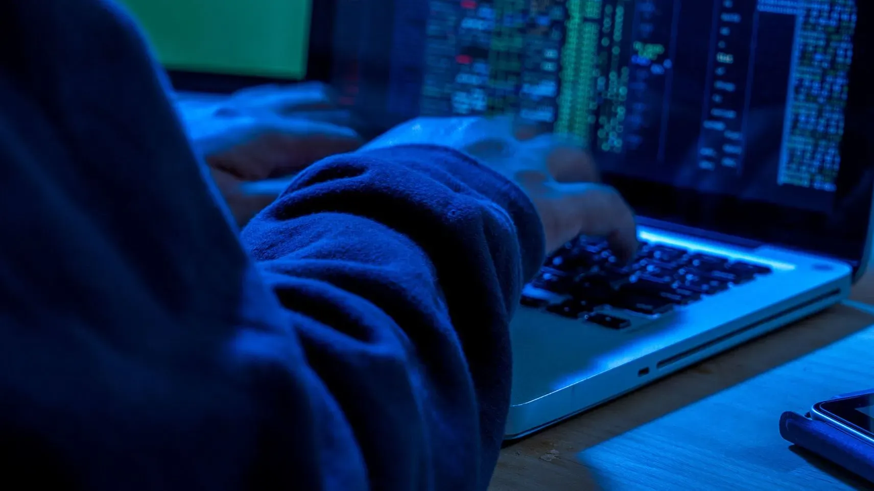 صورة توضح شخصًا يستخدم كمبيوتر محمول في بيئة مظلمة، مع ظهور بيانات وبرامج على الشاشة، تشير إلى أنشطة القرصنة الرقمية.