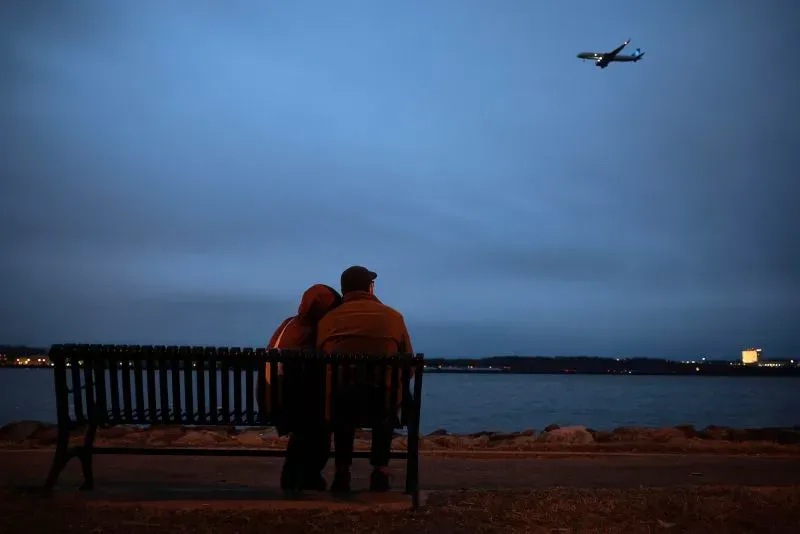 زوجان يجلسان على مقعد بالقرب من الماء، يراقبان طائرة تحلق في السماء المظلمة، تعبير عن التأمل في الحوادث الجوية.