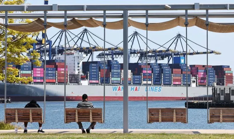 سفينة شحن محملة بالحاويات في ميناء، مع جلوس شخصين على مقاعد خشبية قريبة، تعكس النشاط التجاري المتزايد بين الصين والولايات المتحدة.