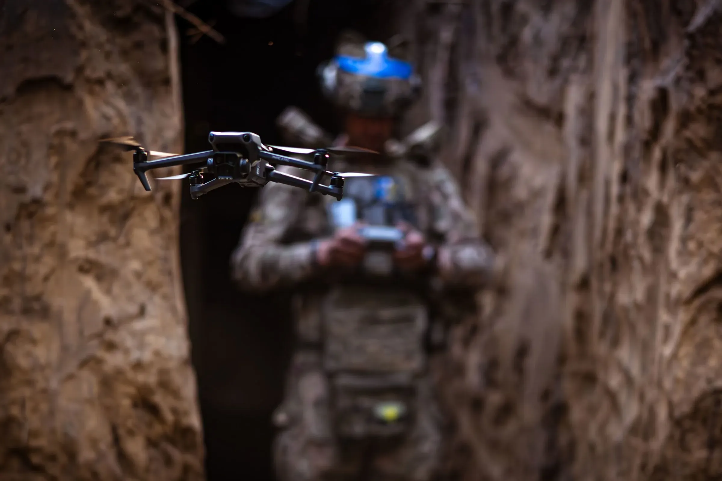 طائرة مسيرة صغيرة تحلق في خندق عسكري، بينما يظهر جندي أمريكي يتحكم بها، مما يعكس استخدام التكنولوجيا الحديثة في ساحة المعركة.