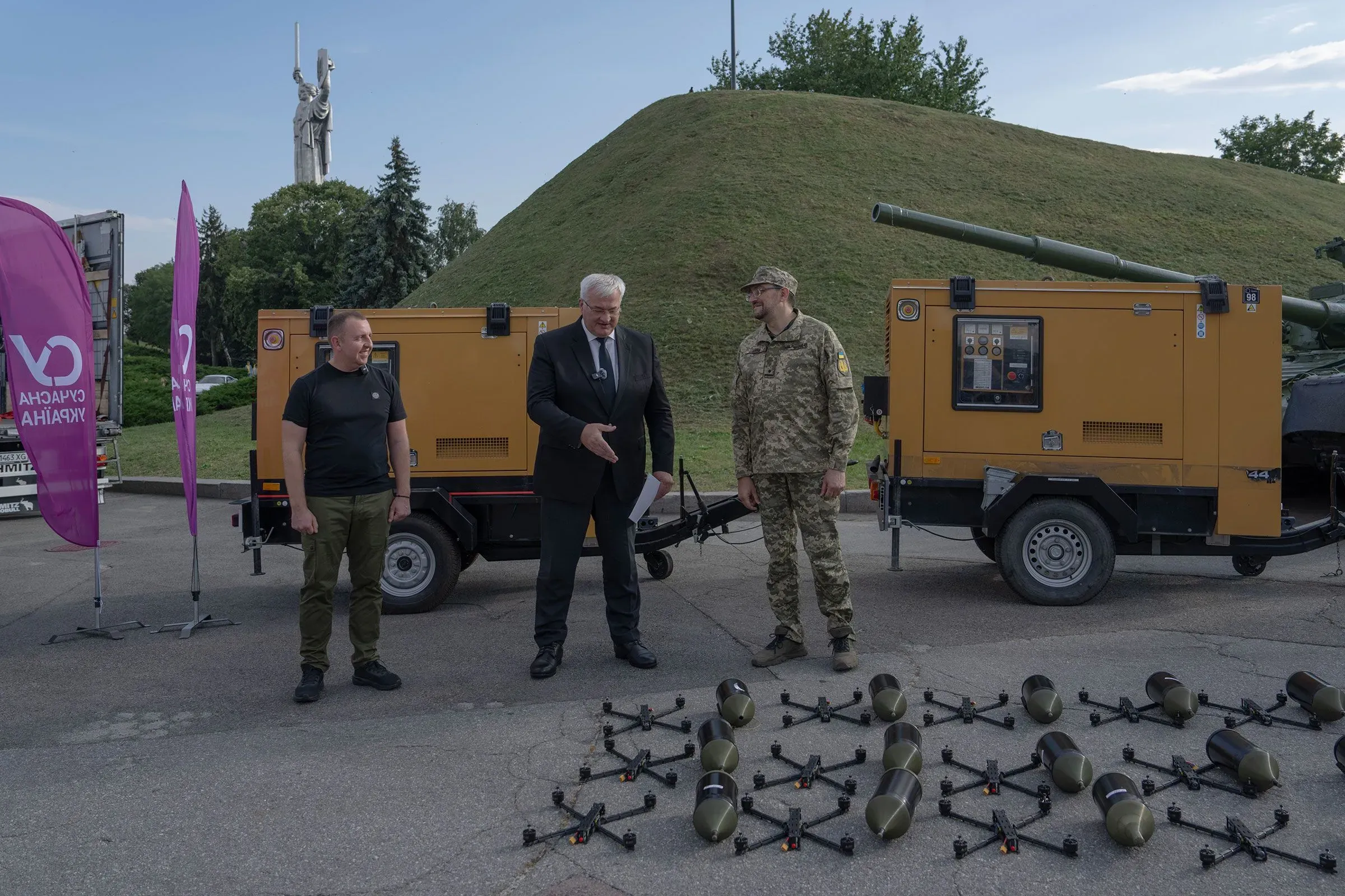 عرض لثلاثة رجال، أحدهم يرتدي زيًا عسكريًا، يتحدثون بجوار طائرات مسيرة وقنابل صغيرة، مع تمثال تاريخي في الخلفية.