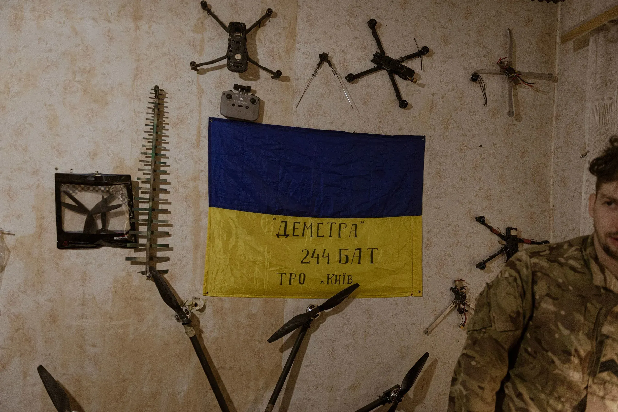جندي أوكراني يقف بجوار علم أوكرانيا في غرفة تحتوي على طائرات درون ومعدات عسكرية، مما يعكس دور التكنولوجيا في النزاع.