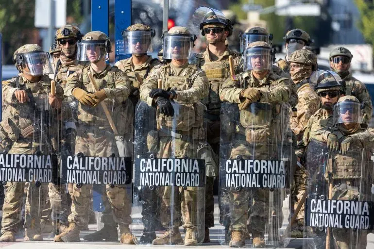 عناصر من الحرس الوطني في كاليفورنيا يرتدون دروعًا واقية ويقفون في صفوف، مما يعكس انتشار القوات خلال الاحتجاجات.