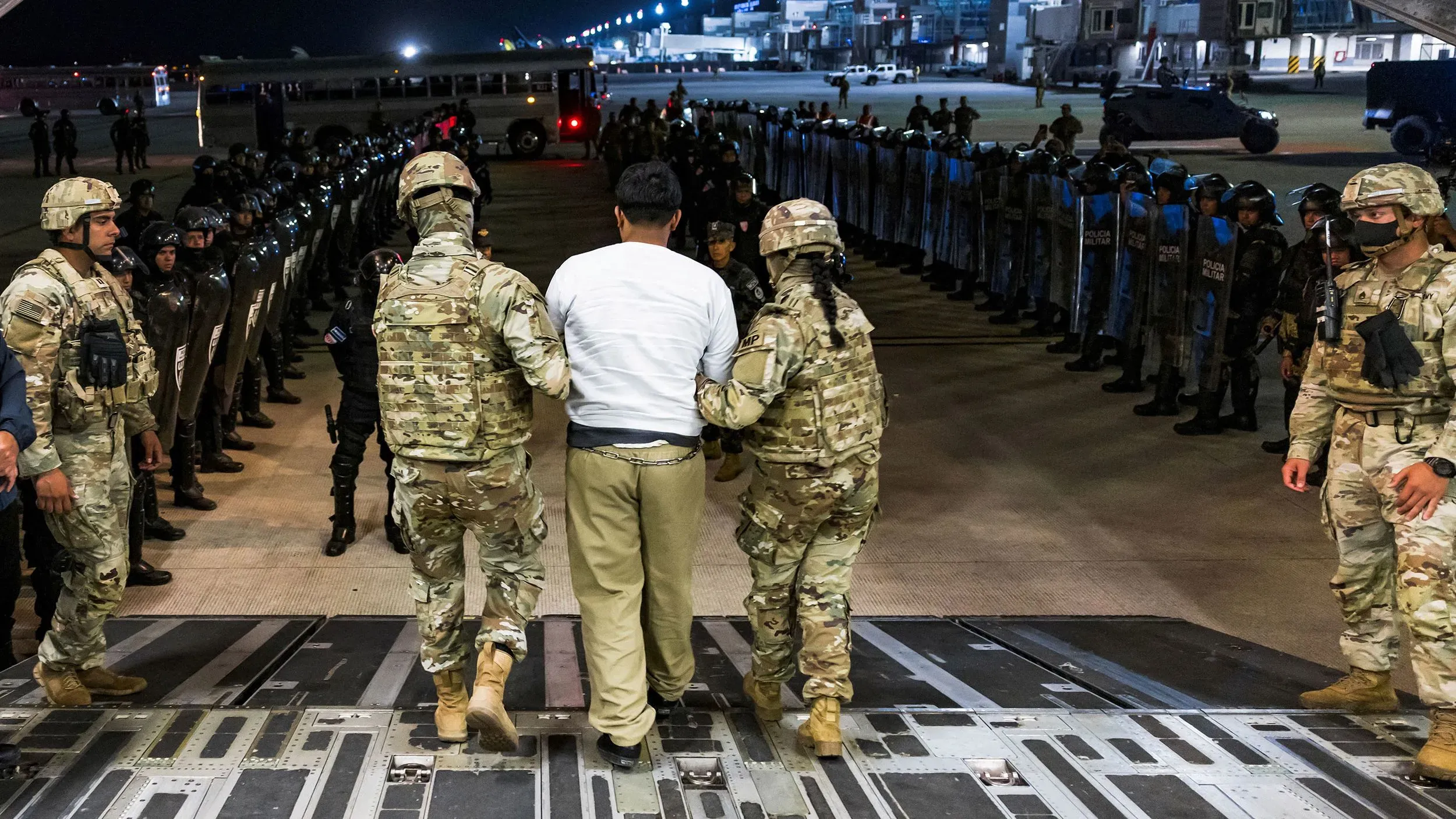 عملية ترحيل مشبوهة لشخص محتجز، بينما يقف أفراد من الشرطة والجيش في صفوف، في مشهد يعكس التوتر الأمني في الولايات المتحدة.