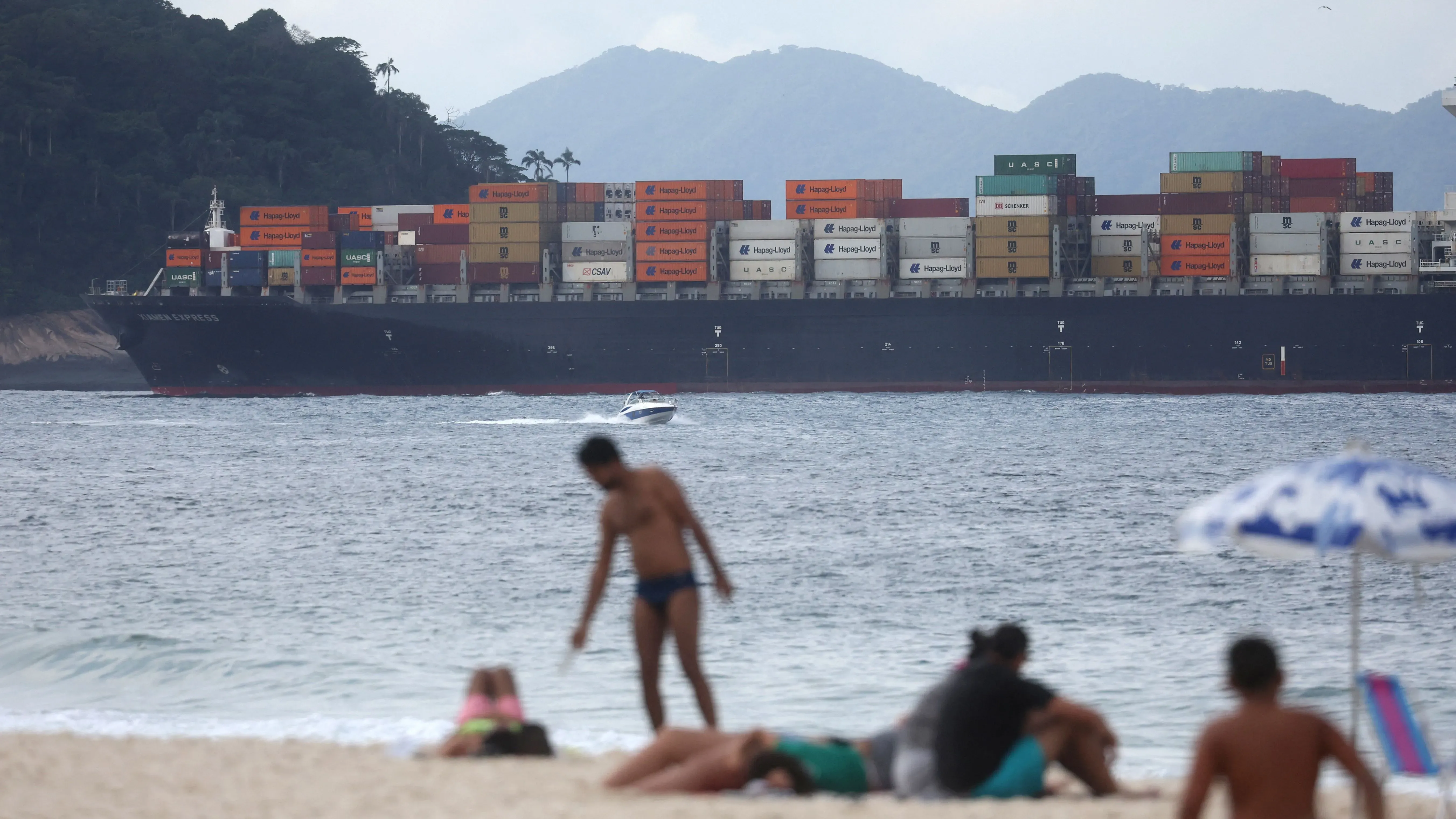 سفينة حاويات كبيرة محملة بالبضائع تبحر قبالة شاطئ مزدحم، حيث يستمتع الناس بأشعة الشمس، مما يبرز أهمية التجارة البحرية في الاقتصاد.