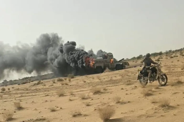 دخان أسود يتصاعد من مركبة محترقة في صحراء النيجر مع رجل على دراجة نارية، يعكس تصاعد العنف والجماعات المسلحة في الساحل.
